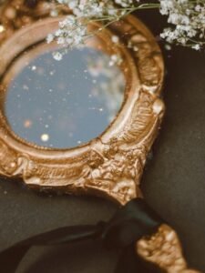 A vintage gold mirror with intricate detailing, tied with a black ribbon and adorned with delicate white flowers, against a dark background.
