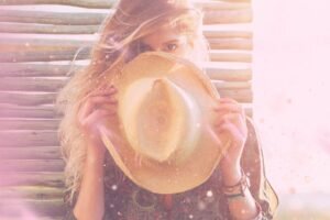 A woman holds a straw hat close to her face, surrounded by soft sunlight and a dreamy, pastel background.