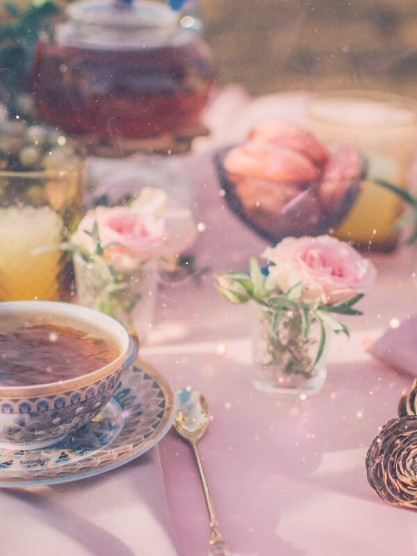 A delicate tea set with a floral cup, surrounded by small pink roses, pastries, and a warm, inviting atmosphere.