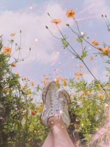 A pair of white sneakers resting on a person's legs surrounded by vibrant orange flowers and a blue sky with soft clouds.