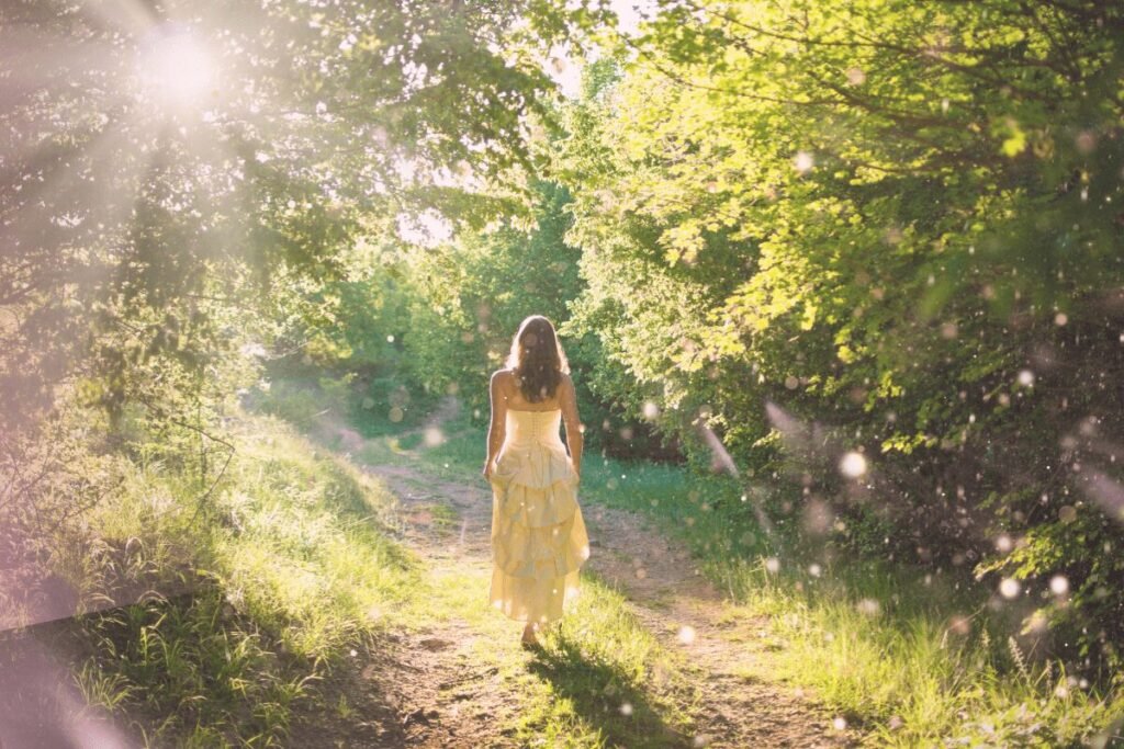 A woman in a flowing yellow dress walks along a sunlit forest path, surrounded by lush greenery and soft, glowing light.